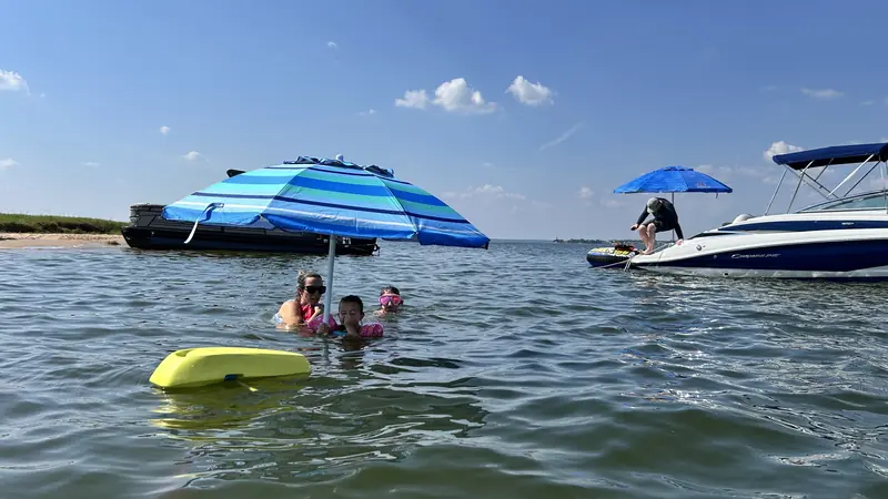Families swimming and playing in Lake Conroe under a blue and teal beach umbrella, with another boat anchored nearby